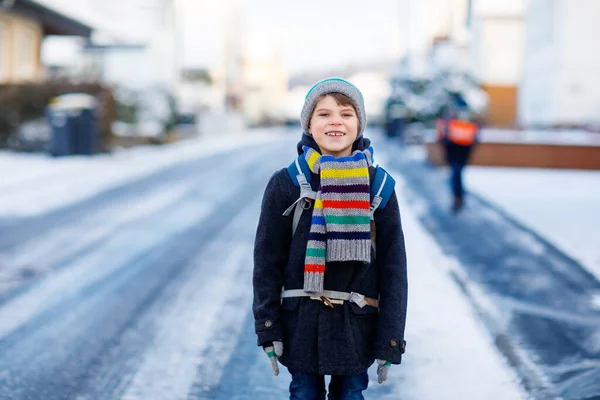 Malý školáček základní třídy, který chodí do školy během sněžení. Šťastné dítě baví a hrát si s prvním sněhem. Student s batohem nebo brašnou v barevných zimních šatech. — Stock fotografie