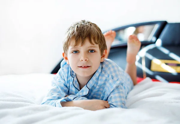 Schattige, gelukkige kleine jongen na het slapen in zijn witte hotelbed in kleurrijke nachtkleding. Schoolkind op familievakantie. — Stockfoto