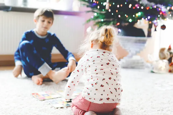 Dvě malé chilren, roztomilé batole dívka a školní kluk hrát spolu karetní hru podle zdobené vánoční stromeček. Šťastní zdraví sourozenci, bratr a sestra se spolu baví. Rodinné oslavy Vánoc — Stock fotografie