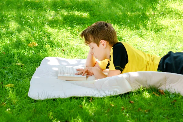 Милый блондин, маленький мальчик читает книжки в саду, на улице. Возбужденный школьник читает громко. Школьник, семья, образование — стоковое фото