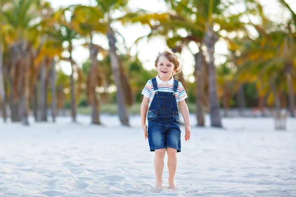 Adorable active little kid boy having fun on tropical beach of island. Happy cute child relaxing, playing, enjoying, running and jumping on sunny warm day near palms and ocean. Active family vacations — Stock Photo, Image