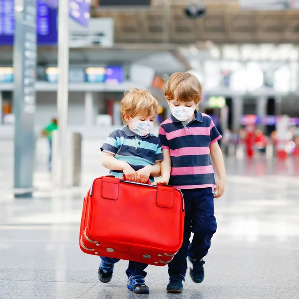 Dois irmãozinhos usam máscara médica e vão de férias com mala no aeroporto, dentro de casa. Irmãos gêmeos felizes caminhando para o check-in ou embarque para o voo durante a pandemia do vírus corona — Fotografia de Stock