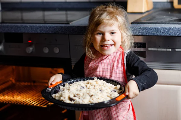 Cute little toddler girl baking apple pie at home. Happy smiling child helping with cake cake in domestic kitchen, standing by hot oven. Healthy homemade food. — Stock Photo, Image