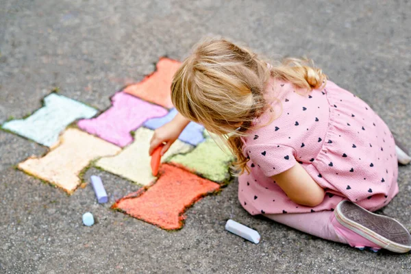 Kleine kleuter meisje schilderen met kleurrijke krijtjes op de grond op de achtertuin. Positief gelukkig peuter kind tekenen en het maken van foto 's. Creatieve buitenactiviteiten in de zomer. — Stockfoto