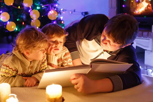 Far och två små barn pojkar läser bok av skorsten, stearinljus och öppen spis. — Stockfoto