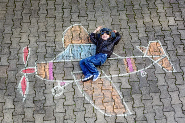 Malý školáček malovat letadlo s barevnými křídy a snít být pilot. Kreativní volný čas pro děti venku v létě. Dítě s helmou a brýlemi. Aktivita pro děti — Stock fotografie