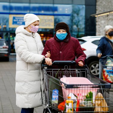 Kıdemli kadın ve tıbbi maskeli sosyal hizmetler görevlisi pandemik koronavirüs hastalığı nedeniyle. Kızı ya da torunu süpermarkette alışveriş yaparken büyükannesine yardım ediyor, market arabasında yiyecek satıyor, açık havada geziyor.