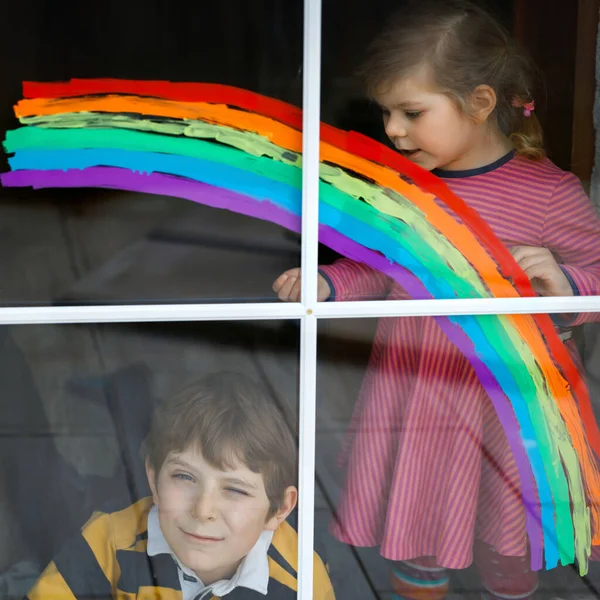 Zwei Kinder, Schuljunge und Kleinkind mit Regenbogen bemalt mit bunten Fensterfarben während der Coronavirus-Quarantäne-Pandemie. Kinder malen Regenbogen mit den Worten Lets all be well — Stockfoto
