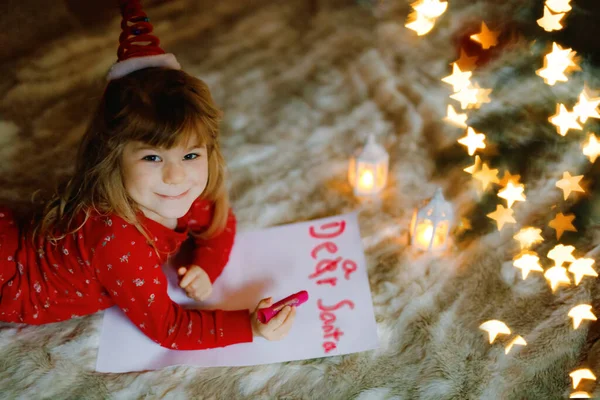 在圣诞树下穿着睡衣的可爱的学龄前小女孩，在家里、室内给圣诞老人写着求职信。传统的基督教节日。快乐蹒跚学步的孩子在圣诞节等待礼物. — 图库照片
