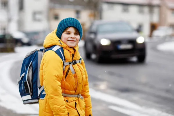 Menino da escola de classe primária a caminhar para a escola durante a queda de neve. Criança feliz se divertindo e brincando com a primeira neve. Estudante com jaqueta amarela e mochila em roupas coloridas de inverno . — Fotografia de Stock