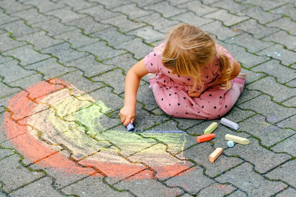 En liten førskolejente som maler regnbue med fargerike kritt på bakken i hagen. Et lykkelig barn som tegner og skaper bilder. Kreativ aktivitet utendørs om sommeren. – stockfoto