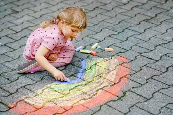 Küçük anaokulu kızı arka bahçede renkli tebeşirlerle gökkuşağını boyuyor. Çocuk resim çizerken ve resim çizerken çok mutlu. Yazın yaratıcı açık hava aktivitesi. — Stok fotoğraf