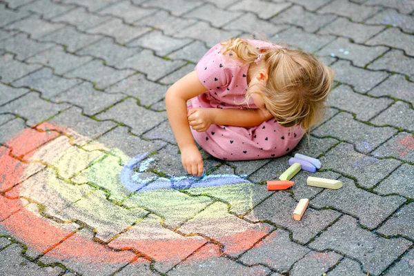 Kleine Vorschulmädchen malen Regenbogen mit bunten Kreiden auf dem Boden im Hinterhof. Positiv glückliches Kleinkind, das zeichnet und Bilder schafft. Kreative Aktivitäten im Sommer. — Stockfoto