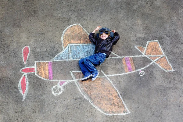 A kis óvodás fiú színes krétákkal festi a repülőt, és arról álmodik, hogy pilóta lesz. Kreatív szabadidő gyerekeknek a szabadban nyáron. Gyerek sisakkal és szemüveggel. Tevékenység gyerekeknek — Stock Fotó
