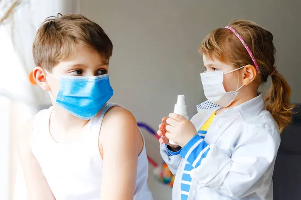 La niña le inyecta al hermano, al chico de la escuela. Niños, hermanos con máscara médica jugando al doctor, sosteniendo jeringa con vacuna. Concepto de vacunación covid contra el Coronavirus. Los niños juegan papel juego —  Fotos de Stock