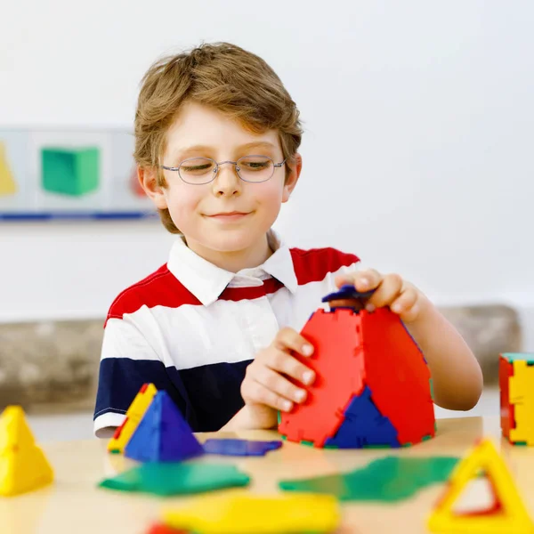 Ragazzino con gli occhiali che gioca con il kit di elementi di plastica lolorful nella scuola o nella scuola materna. Felice bambino che costruisce e crea figure geometriche, impara matematica e geometria. — Foto Stock