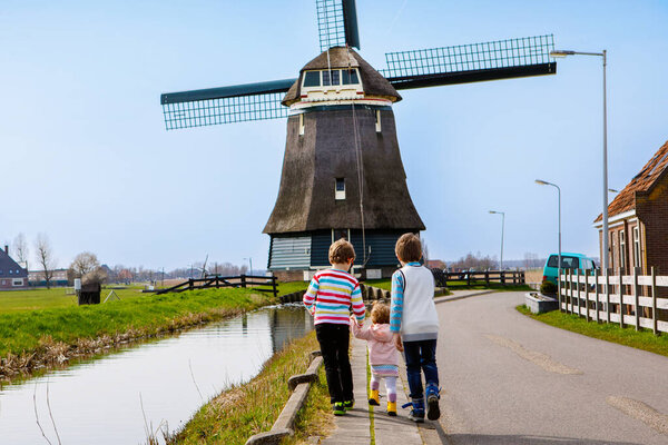 Two school kids boys and little baby girl walking on street with big windmill in Netherlands. Family of three children making vacations in Holland. Happy siblings holding hands.