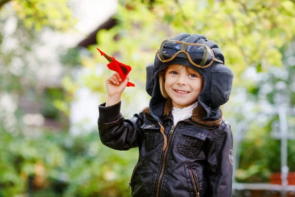 Niño feliz niño en casco piloto y uniforme jugando con el avión de juguete rojo. Sonriente niño preescolar soñando y divirtiéndose. Educación, profesión, concepto de sueño —  Fotos de Stock