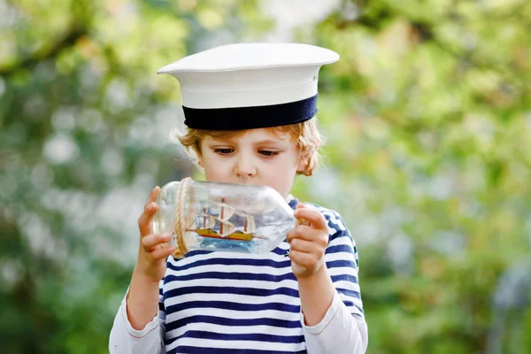 Glad liten pojke i sjöman capitain hatt och uniform leker med segelbåt fartyg. Leende förskolebarn drömmer och har roligt. Utbildning, yrke, drömbegrepp — Stockfoto