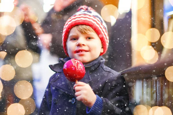 可爱的小男孩在德国圣诞市场上吃甜红苹果。穿着冬衣的快乐孩子,背景色明亮.小孩看着摄像机。家庭、传统、假日概念. — 图库照片
