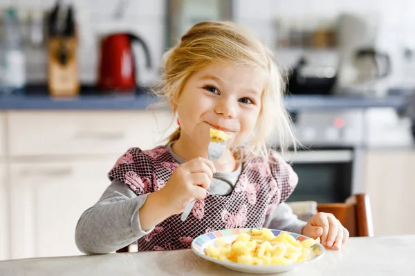 Lief peutermeisje dat gezonde gebakken aardappelen eet als lunch. Schattig vrolijk baby kind in kleurrijke kleding zittend in de keuken van huis, kinderopvang of kinderkamer. Kind eet groenten. — Stockfoto