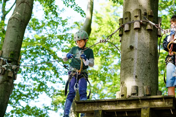 Two children in forest adventure park. Kids boys in helmet climbs on high rope trail. Agility skills and climbing outdoor amusement center for children. Outdoors activity for kid and families. — Stock Photo, Image