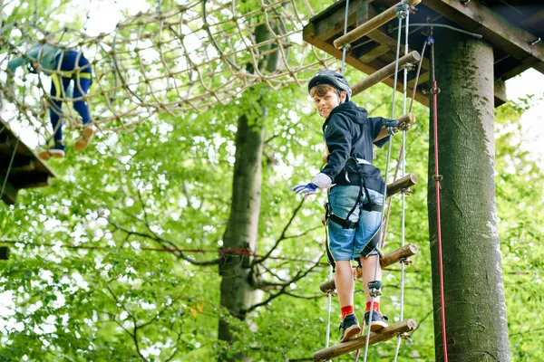 Two children in forest adventure park. Kids boys in helmet climbs on high rope trail. Agility skills and climbing outdoor amusement center for children. Outdoors activity for kid and families. — Stock Photo, Image