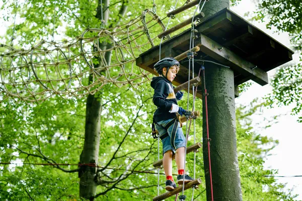 Skolpojke i skogen äventyrspark. Acitve barn, unge i hjälm klättrar på hög rep spår. Agility färdigheter och klättring utomhus nöjescentrum för barn. Friluftsaktivitet för barn och familjer. — Stockfoto