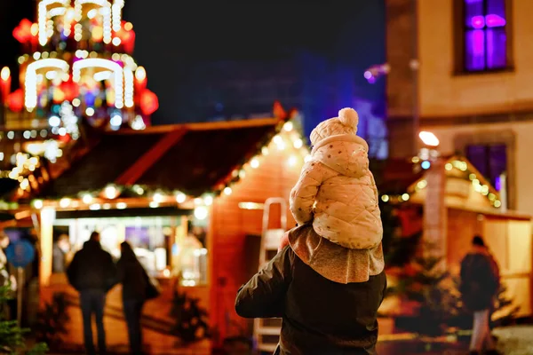 ドイツのクリスマスマーケットで父の肩に座っている小さな就学前の女の子。伝統的な装飾されたピラミッドを観察する幸せな幼児の子供と男。幸せな家族、絆、愛。家族のクリスマスの時間. — ストック写真