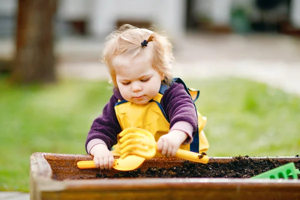 Cute adorable toddler girl playing with sand and shovel on spring day. Baby child wearing yellow boots and mud rain puddle pants. Happy girl planting vegetables in spring. — Stock Photo, Image