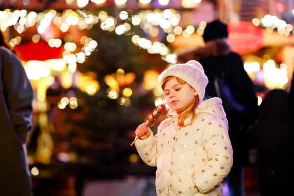 Little preschool girl, cute child eating fruits, strawberries covered with chocolate with decoration and lights on background. Happy child on Christmas market in Germany. — Stock Photo, Image