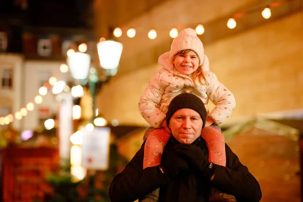 ドイツのクリスマスマーケットで父の肩に座っている小さな就学前の女の子。伝統的な装飾されたピラミッドを観察する幸せな幼児の子供と男。幸せな家族、絆、愛。家族のクリスマスの時間. — ストック写真