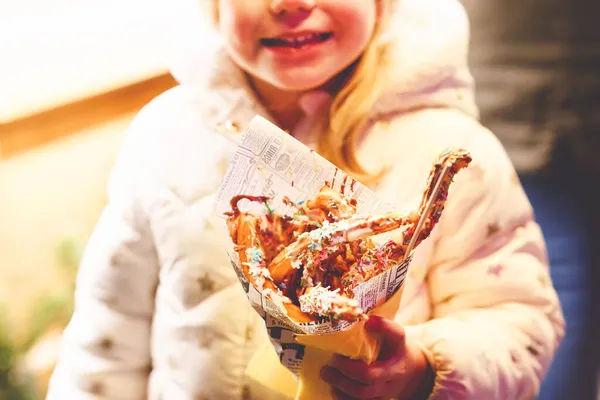 Klein kleuter meisje, schattig kind eten churros snoep bedekt met chocolade met decoratie en verlichting op de achtergrond. Gelukkig kind op kerstmarkt in Duitsland. — Stockfoto