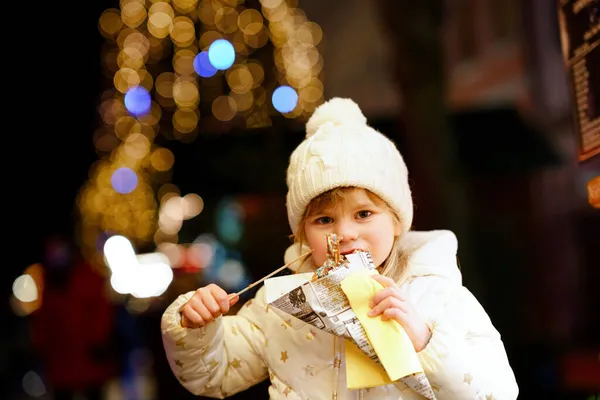 小さな就学前の女の子、かわいい子供が背景に装飾や照明でチョコレートで覆われたチュロスのお菓子を食べています。ドイツのクリスマスマーケットで幸せな子供. — ストック写真
