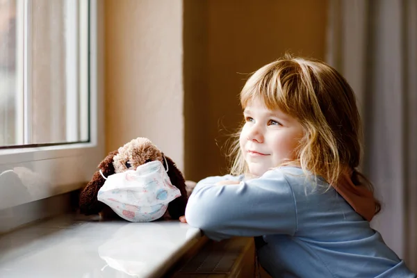 かわいい幼児の女の子が窓のそばに座って外を見ています。夢の子供は幸せな感じ柔らかい犬のおもちゃに医療用マスクを置きます。コロナウイルスのパンデミック時のロックダウンと隔離の概念。孤独な子. — ストック写真