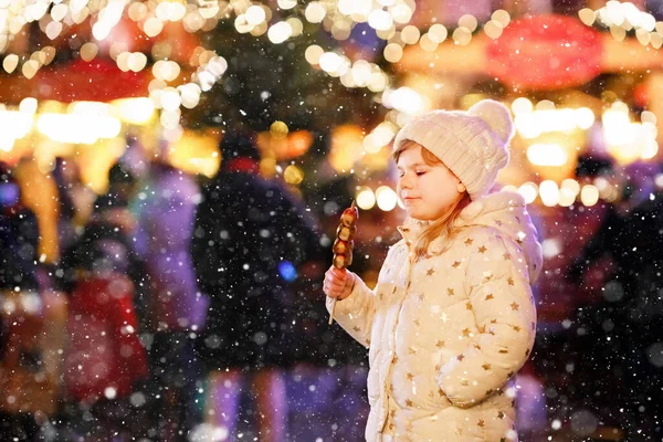 Kleine Vorschulmädchen, niedliches Kind, das Früchte isst, Erdbeeren mit Schokolade bedeckt mit Dekoration und Lichtern auf dem Hintergrund. Glückskind auf dem Weihnachtsmarkt in Deutschland. — Stockfoto