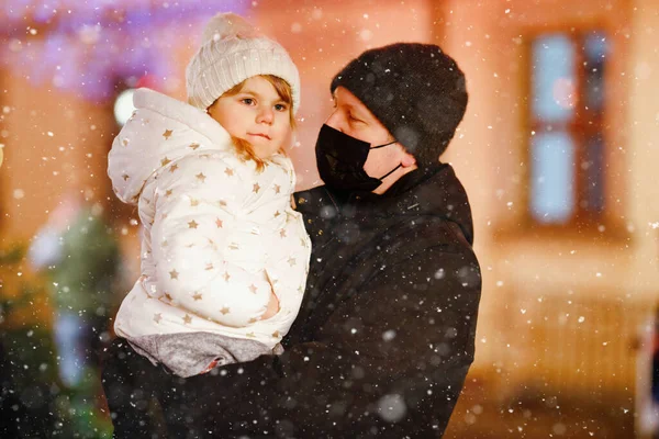 Malá školačka sedící na otcově rameni na vánočním trhu v Německu. Šťastné batole dítě a muž s lékařskou maskou kvůli pandemii viru korony. Rodina, sblížení, láska. Rodinný vánoční čas. — Stock fotografie