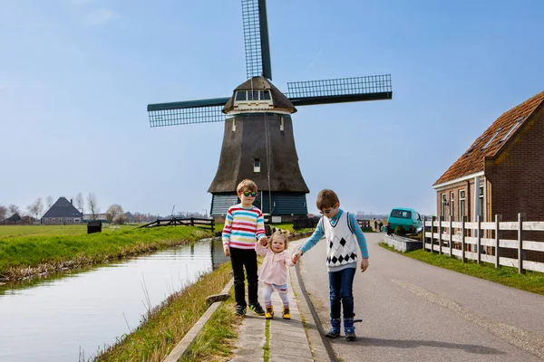 Due ragazzi della scuola e una bambina che camminano per strada con un grande mulino a vento in Olanda. Famiglia di tre bambini in vacanza in Olanda. Fratelli felici che si tengono per mano. — Foto Stock