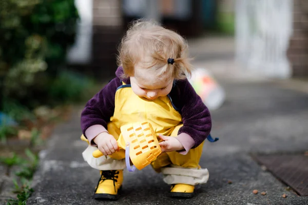 Menina adorável bonito criança brincando com areia e pá no dia de primavera. Criança usando botas amarelas e calça de chuva de lama. Menina feliz plantando legumes na primavera. — Fotografia de Stock