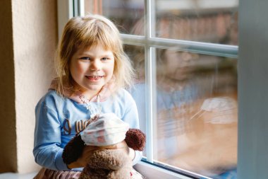 Pencere kenarında oturan ve dışarı bakan tatlı bir kız. Rüya gören çocuk mutlu hissederek yumuşak köpek oyuncağına tıbbi maske taktı. Corona virüsü salgını sırasında tecrit ve tecrit kavramı. Yalnız çocuk..