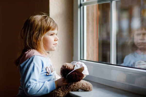 かわいい幼児の女の子が窓のそばに座って外を見ています。夢の子供は幸せな感じ柔らかい犬のおもちゃに医療用マスクを置きます。コロナウイルスのパンデミック時のロックダウンと隔離の概念。孤独な子. — ストック写真