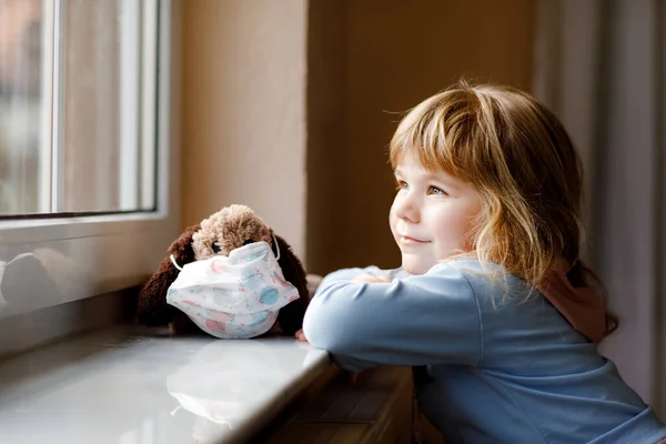 Милая девочка, сидящая у окна и выглядывающая. Мечтающий ребенок надел медицинскую маску на мягкую собачью игрушку, чувствуя себя счастливым. Концепция изоляции и изоляции во время пандемии коронного вируса. Одинокий ребенок. — стоковое фото