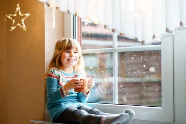 学龄前小女孩拿着一杯热巧克力和玛哈蜜糖。快乐的孩子在冬天开着圣诞灯，在窗边喝甜可可。舒适的圣诞家庭庆祝活动. — 图库照片