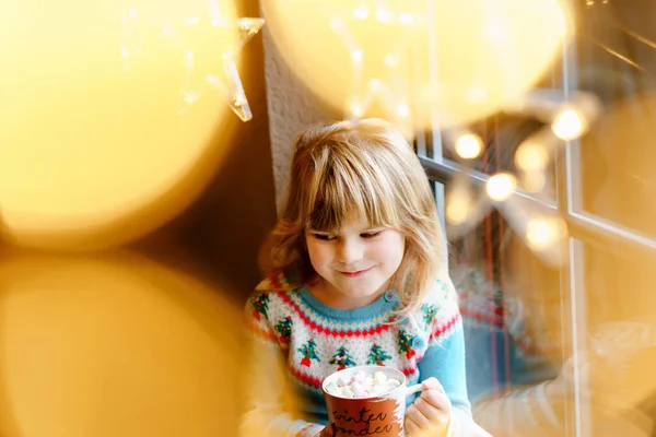 Little preschool girl holding cup with hot chocolate with marhsmallows. Happy child drinking sweet cocoa by window with Christmas lights in winter. Cozy family celebration of xmas. — Stock Photo, Image