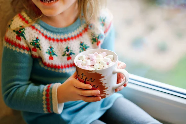 学龄前小女孩拿着一杯热巧克力和玛哈蜜糖。快乐的孩子在冬天开着圣诞灯，在窗边喝甜可可。舒适的圣诞家庭庆祝活动. — 图库照片