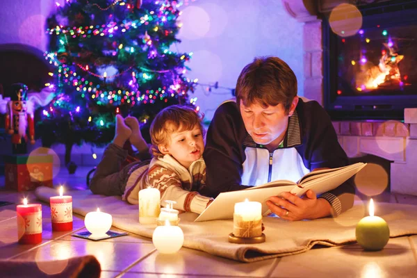 Vater und süßer kleiner Junge lesen Buch am Kamin, Kerzen und Kamin. — Stockfoto
