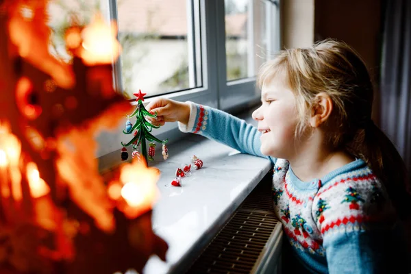 Niña pequeña sentada junto a la ventana y decorando un pequeño árbol de Navidad de vidrio con pequeños juguetes de Navidad. Feliz niño sano celebrar la fiesta tradicional familiar. Adorable bebé.. — Foto de Stock