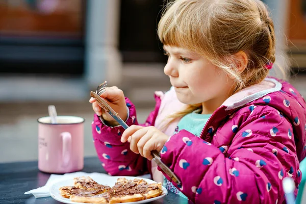 屋外カフェレストランに座ってかわいい就学前の女の子。幸せな健康な子供は甘いワッフルかクレープを食べ、ナイフを使用し、晴れた日にフォークを学ぶ。ホットチョコレートを飲む子供 — ストック写真