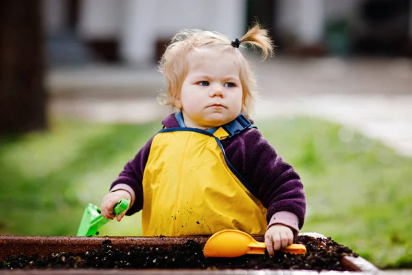 Niedliche entzückende Kleinkind Mädchen spielen mit Sand und Schaufel am Frühlingstag. Baby in gelben Stiefeln und Schlammpfützenhosen. Glückliches Mädchen pflanzt Gemüse im Frühling. — Stockfoto