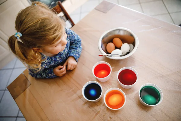 Excited little toddler girl coloring eggs for Easter. Cute happy child looking surprised at colorful colored eggs, celebrating holiday with family. From above, unrecognized face. — Stock Photo, Image